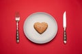 Heartbreaker concept image: heart shaped cookie on plate with fork and knife over red background