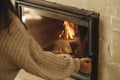 Heating house in winter with wood burning stove. Woman putting firewood into burning fireplace in rustic room in farmhouse.