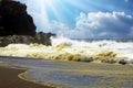 Heavy violent surf waves breaking on secluded black lava sand beach and rocks, sea spray and foam, sun rays clouds backlight  - Royalty Free Stock Photo