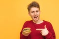 ÃÂ¡heerful young man stands on a yellow background with a burger in his hand, shows his finger on the burger and looks at the