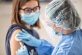 Helathcare employee wearing gloves, protective shield and a cap applies vaccine from syringe into the arm of a young patient