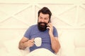 Hello dear. Bearded man using mobile technology in bed. Handsome guy talking on phone and drinking coffee at home Royalty Free Stock Photo