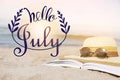 Hello July. Open book, sunglasses and hat on sandy beach near sea