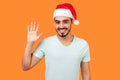 Hello! Portrait of positive handsome bearded man in santa hat and casual white t-shirt waving raised hand and saying hi to camera Royalty Free Stock Photo