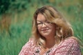 Hello, the sun! A young fat woman with lush hair and glasses sits on a sunlit glade and squintes