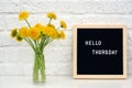 Hello Thursday words on black letter board and bouquet of yellow dandelions flowers on table against white brick wall. Concept