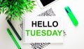 HELLO TUESDAY written in black and green type on white paper near green markers, a smartphone and a green plant on a white
