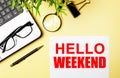 HELLO WEEKEND written on white paper near a computer, magnifier, glasses, pens on a yellow background. Motivational concept. Flat Royalty Free Stock Photo