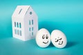 The hen eggs with painted faces. The concept. She leaned against near a white paper house on a blue background. Royalty Free Stock Photo