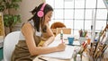 At her art studio, a young, beautiful hispanic woman artist is seriously concentrated, drawing in her notebook, headphones on, Royalty Free Stock Photo