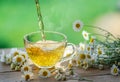 Herbal chamomile tea and chamomile flowers near teapot and tea glass on wooden table. Countryside background