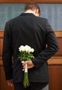 Hes a man of mystery. A man wearing a suit holding a bunch of white roses behind his back.