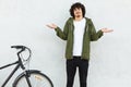 Hesitant curly man shruggs shoulders in bewilderment, dressed in fashionable green anorak, stands near bicycle, isolated over