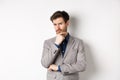 Hesitant young businessman in suit looking skeptical at camera, thinking and making decision, standing against white Royalty Free Stock Photo