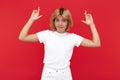 Hesitating young blonde woman in casual white outfit holding hands with fingers crossed in hope over red background.