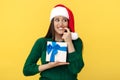 Hesitating young brunette woman wear santa claus hat and green pullover holding gift box over yellow background.