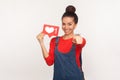 Hey you, like and follow interesting content! Portrait of positive stylish girl in denim overalls holding social media heart icon