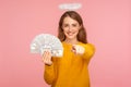 Hey you, make money! Portrait of positive angelic ginger girl with halo over head holding fan of dollar bills and pointing finger