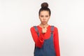 Hey you! Portrait of bossy dissatisfied angry girl with hair bun in denim overalls pointing finger to camera, blaming