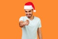 Hey you! Portrait of young joyful bearded man in santa hat and casual white t-shirt standing, pointing finger and looking at Royalty Free Stock Photo