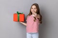 Little girl wearing striped T-shirt holding present box and pointing finger to camera. Royalty Free Stock Photo