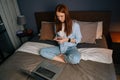 High-angle shot of beautiful redhead young woman typing message on mobile phone sitting on bed