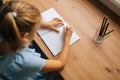 High-angle side view of unrecognizable primary little child girl drawing with pen making picture sitting at table in Royalty Free Stock Photo