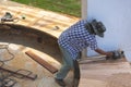 Asian carpenter using electric planer to shaving curved wooden bench surface at outdoor workshop