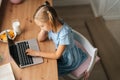 High-angle view of bored elementary child girl using typing laptop keyboard sitting at home table with snack by window. Royalty Free Stock Photo