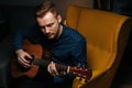 Close-up portrait of handsome guitarist singer male playing acoustic guitar sitting on armchair in dark living room. Royalty Free Stock Photo