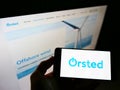 High angle view of person holding cellphone with logo of Danish power company ÃËrsted AS on screen in front of webpage.
