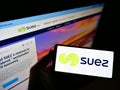 High angle view of person holding cellphone with logo of French utility company Suez SA on screen in front of website.