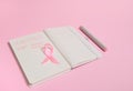 Top view of a pink ribbon, on opened diary with inscriptions reminding of a medical check-up. October 1st, World Breast Cancer Day