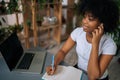 High-angle view of pretty curly black female in earphones holding pen in hand sitting at desk with laptop, paper book Royalty Free Stock Photo