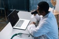 High-angle view of tired black male doctor wearing white coat uniform using working on laptop computer sitting at desk Royalty Free Stock Photo