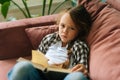 High-angle view of unhappy adorable child kid girl reading paper book lying on soft couch at home looking at camera. Royalty Free Stock Photo