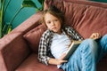 High-angle view of unhappy adorable child kid girl reading paper book lying on soft couch at home looking at camera. Royalty Free Stock Photo