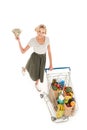 high angle view of young woman holding dollar banknotes and smiling at camera while standing with shopping trolley full of Royalty Free Stock Photo