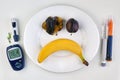 Banana and crushed plums in the form of a horror emoticon on a white plate and insulin syringe pens and a glucometer as