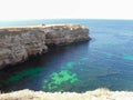 A high long rock that goes into the sea and a car and a man on top of the cliff. Sunny day, the sky and the clear turquoise water