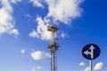 The high mast or pole with spotlights and a traffic sign `go straight or left turn` against a blue sky with white clouds, low a Royalty Free Stock Photo