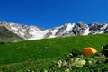 High snow mountain range above a small orange tent in green valley