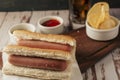 High view of a pair of plain hotdogs with pots of mustard and ketchup and some potato chips on a rustic wooden tray. Horizontal