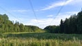 A high-voltage power line on metal poles runs along a grassy clearing in a mixed forest. Sunny summer weather and a blue sky with Royalty Free Stock Photo