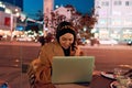 A hijab woman using laptop and smartphone at a modern coffe in the nighttime cityscape, reflecting the harmonious blend