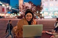 A hijab woman using laptop and smartphone at a modern coffe in the nighttime cityscape, reflecting the harmonious blend