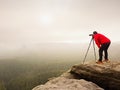 Hiker with camera on tripod takes picture from rocky summit. Alone photographer on summit