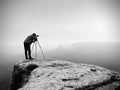 Hiker with camera on tripod takes picture from rocky summit. Alone photographer on summit