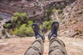 A hiker dangles his feet over the edge of a cliff looking down into a desert canyon.