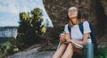 Hiker girl with backpack and ÃÂup of drink in her hand sitting in mountains and plans trip route on map, female person in glasses